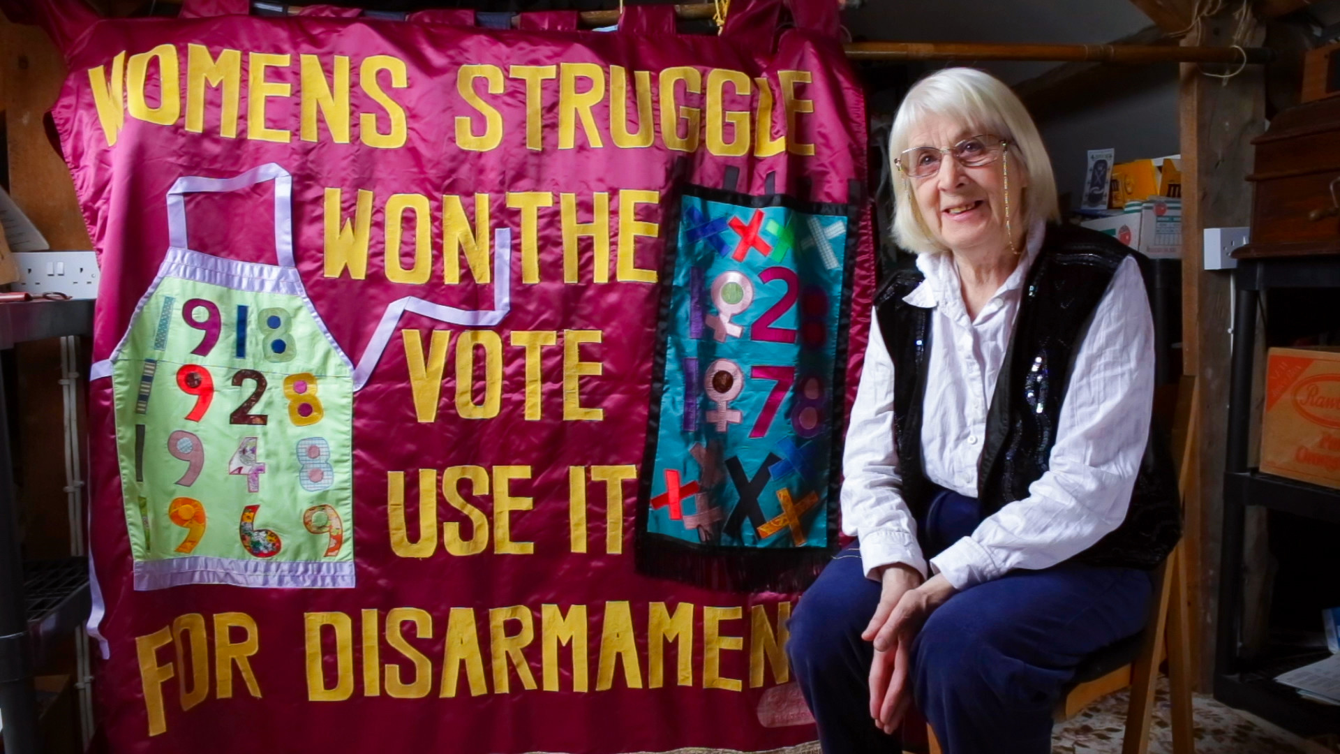 Thalia, an old white woman, sit in front of a larger banner she made